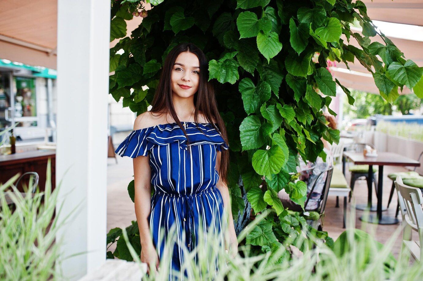 Woman in blue striped dress standing next to lush green leaves in an outdoor café.