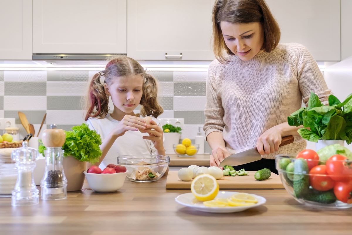 Mother and child cooking together at home in kitchen. Healthy eating, mother teaches daughter to cook, parent child communication. Woman cutting cucumber, girl peeled boiled egg.